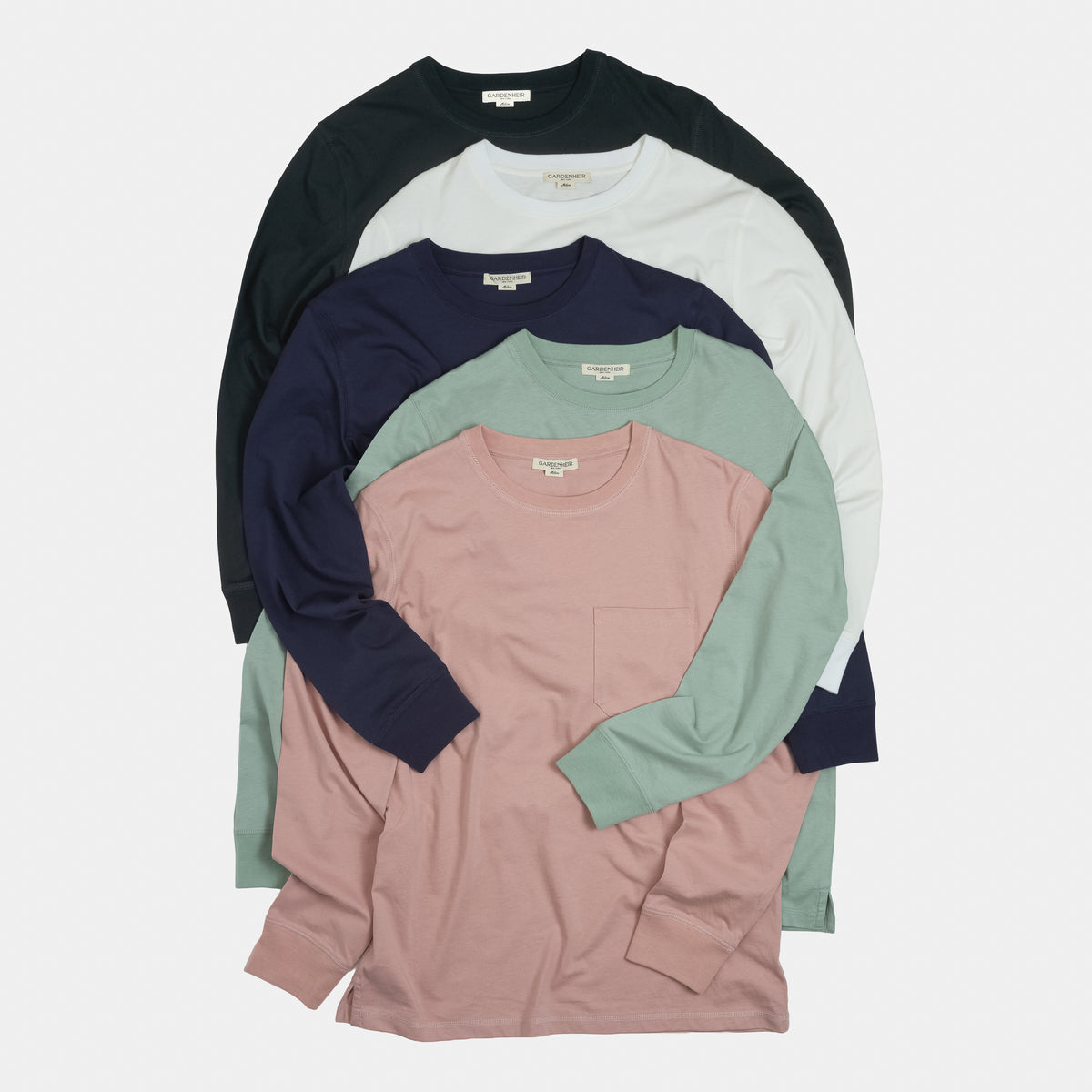 A stack of five long-sleeve crewneck pocket shirts in different colors: black, off-white, navy, sage green, and dusty pink, arranged neatly on a light gray background.