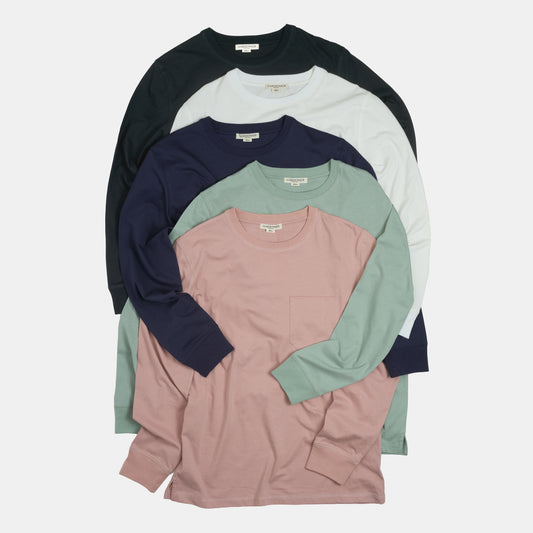 A stack of five long-sleeve crewneck pocket shirts in different colors: black, off-white, navy, sage green, and dusty pink, arranged neatly on a light gray background.